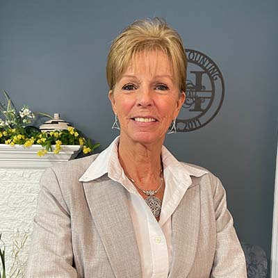 Tina Hall in a gray suit with a white blouse, standing indoors with a decorative background.